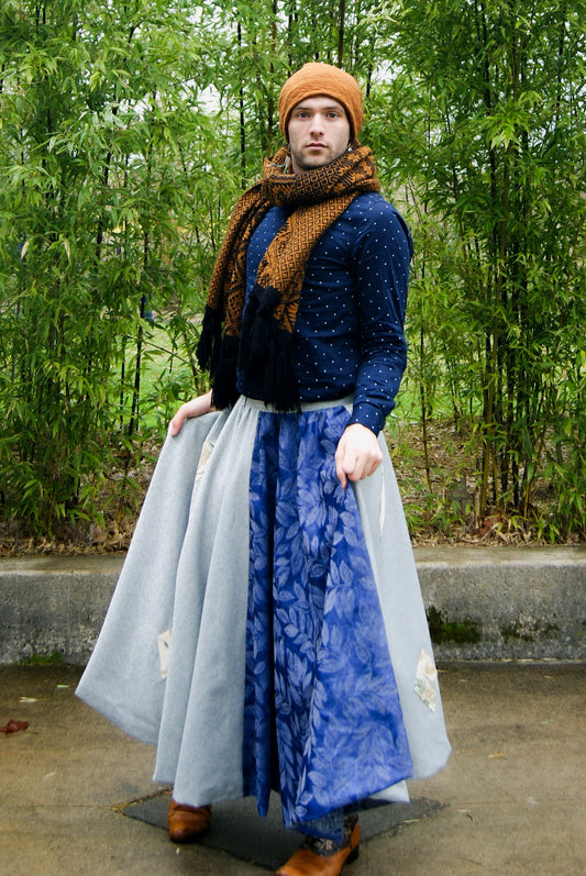 Image shows a tall, slightly bearded non binary person, wearing an orange-brown toque, a blue dress shirt, fringe earrings in blue, and a four paneled circle skirt made from repurposed curtain panels. They are facing the camera with their arms by their sides, holding their skirt and about to do a big spin around