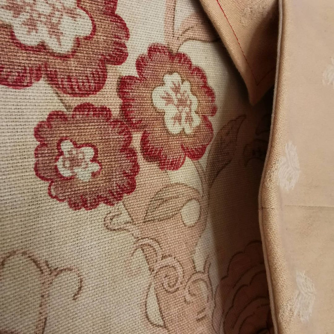 Image is a close up of a medium weight woven cotton fabric, with pastel tones of pink and beige. There are marigolds printed on the fabric in a deep red, and there is a piece of a peach coloured fabric laying over the main material. The stitching is red