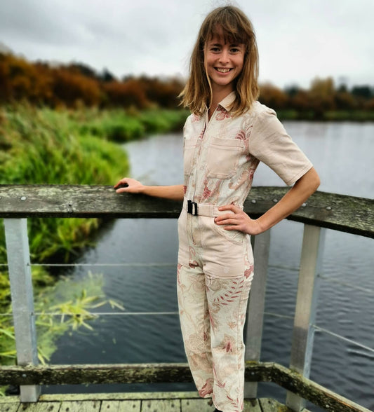 Image is of a model, standing at a lookout point at Swan Lake in Victoria BC. She is wearing a shortsleeved jumpsuit made from a medium weight woven cotton fabric. The colour of the fabric is a mix of pastels, with subtle prints of leaves and flowers in a deep red. The garment has lots of pockets, and a fabric belt with buckle closure. The model is staring into the camera and smiling with one hand on her hip, and one on a wooden railing. Behind her is the lake, and long grasses