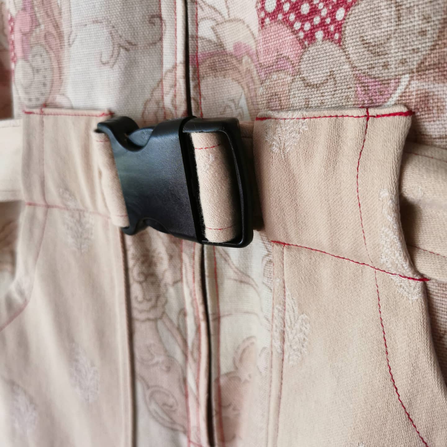 Image centers on a black buckle that is closed over a stitched zipper. There is a belt through the buckle, made from a peach fabric, which is also visible on either side of the zipper, as pockets. The fabric is primarily pastels and pink but there is an ornate design printed in deep red which is visible at the corner of the image.