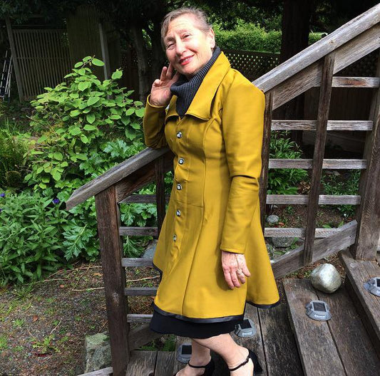 Image is of a woman with cropped short hair, standing on a wooden staircase in the backyard of a home in Victoria BC. She has an elbow rested on the railing with her hand on her cheek, smiling towards the camera. She is wearing a handmade mustard yellow Fall coat with a fitted waist and flared hem. The coat is buttoned up, and there is a turtleneck peeking out at the neckline.