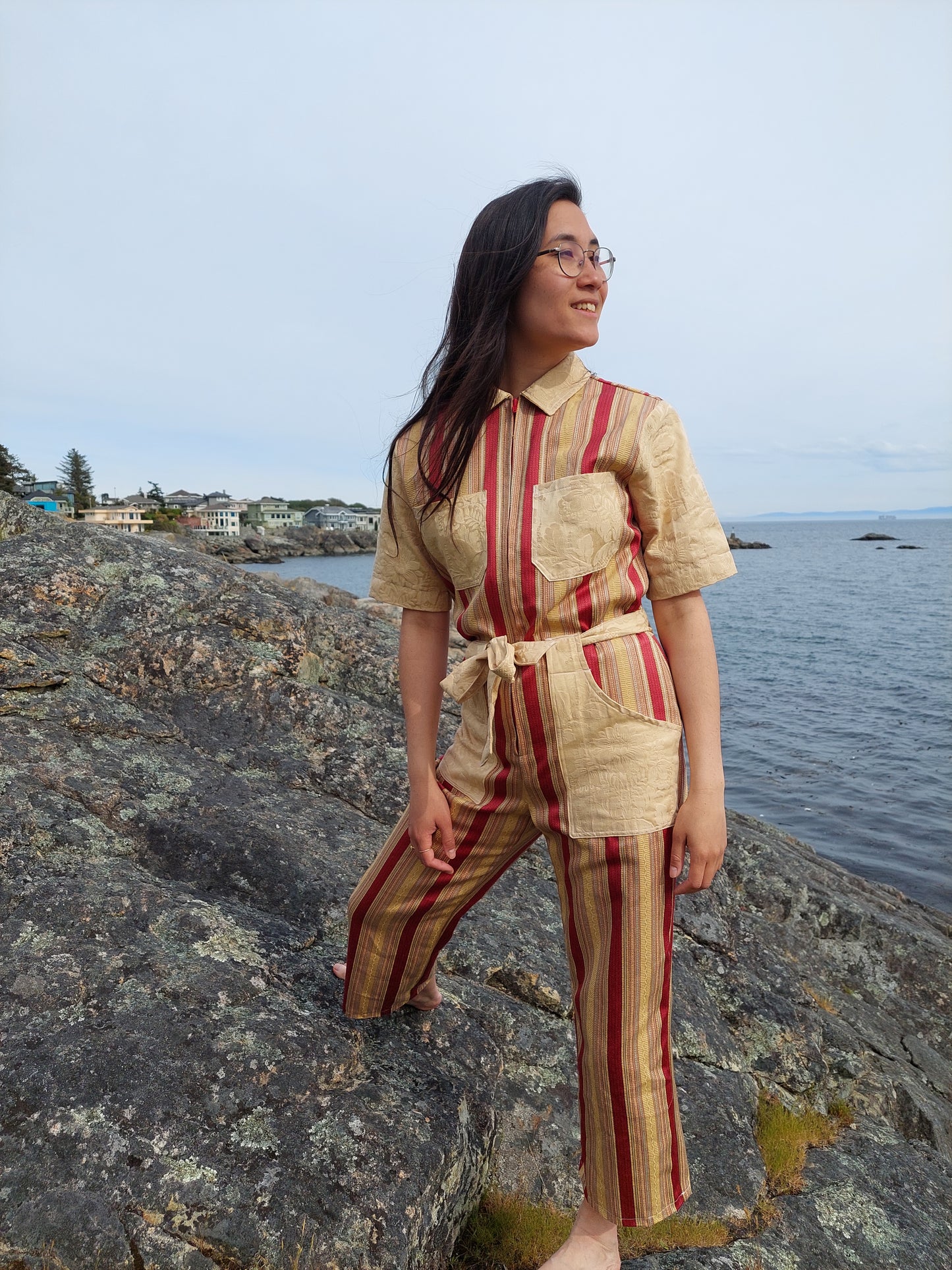 Model stands on the rocks at Saxe point, in Victoria BC. She looks towards the ocean, and her hair falls over the striped gold and red handmade jumpsuit she wears. She is smiling and looks peaceful