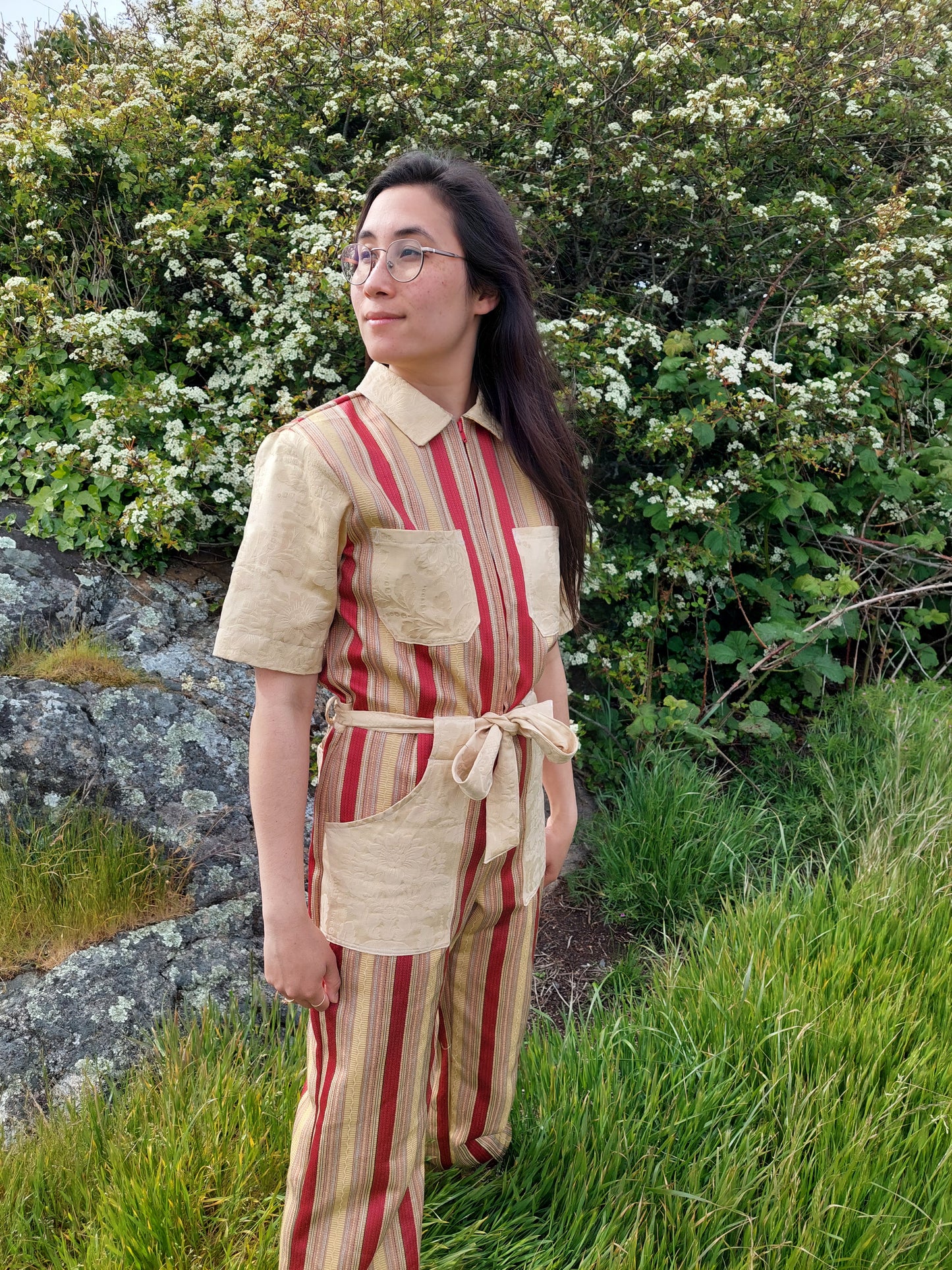 Model stands on in tall grass, in front of flowering bushes at Saxe point, in Victoria BC. She looks towards the ocean, and her hair falls over the striped gold and red handmade jumpsuit she wears. She is smiling and looks peaceful