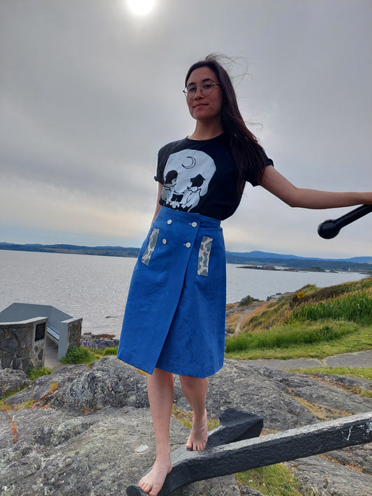 Model is standing facing the camer, at Saxe Point in Victoria BC, they are standing on an anchor which is displayed on the rocks and they are holding onto a piece of the anchor for stability. They are wearing a cobalt blue A line skirt with 4 white buttons on the front and two small pocket details on the front in a chalky blue. Behind the model, the day has turned slightly stormy, with darker grey clouds gathering a bit. The models hair is being pulled by the wind, but they don't seem affected