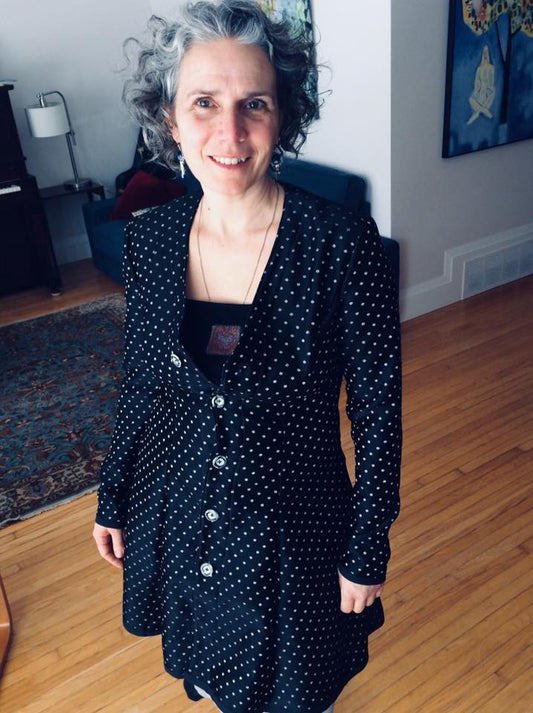 Image is of a woman standing facing the camera in a well lit living room. She has cropped black and silver hair, and is smiling. She wears a fitted black coat, with small polks dots tightly packed onto the fabric. The coat is buttoned up with 5 four hole buttons