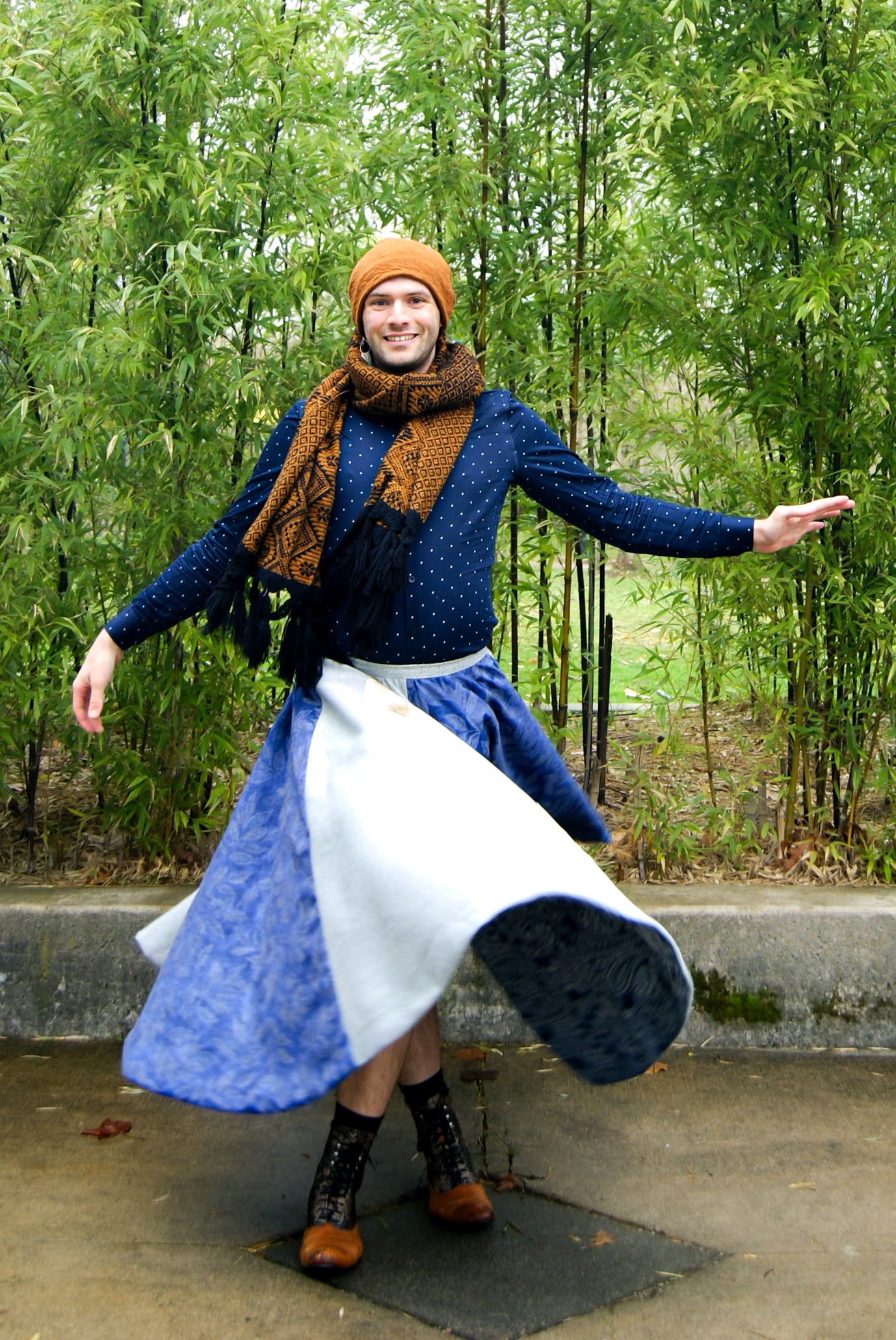 Image shows a tall, slightly bearded non binary person, wearing an orange-brown toque, a blue dress shirt, fringe earrings in blue, and a four paneled circle skirt made from repurposed curtain panels. They have just stopped spinning around and the skirt is catching up to them and slowly returning to a falling position. They are facing the camera with one arm by their side, and the other slightly lifted, mid spin