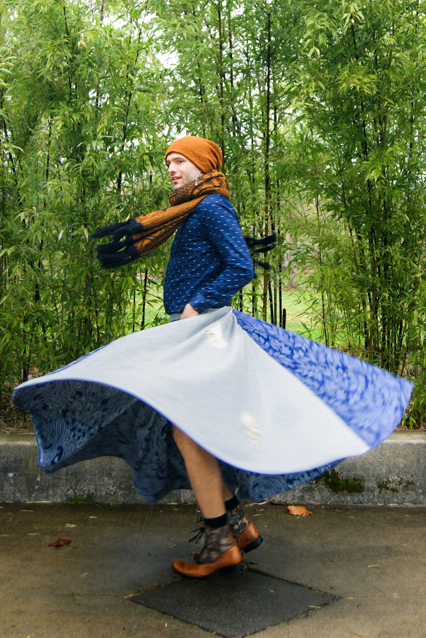 Image shows a tall, slightly bearded non binary person, wearing an orange-brown toque, a blue dress shirt, fringe earrings in blue, and a four paneled circle skirt made from repurposed curtain panels. They are spinning around and the skirt is in full movement, and they are facing the side with their arms by their sides, mid spin