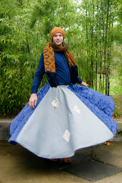 Image shows a tall, slightly bearded non binary person, wearing an orange-brown toque, a blue dress shirt, fringe earrings in blue, and a four paneled circle skirt made from repurposed curtain panels. They are spinning around and the skirt is in full movement, and they are facing the camera with their arms by their sides, mid spin