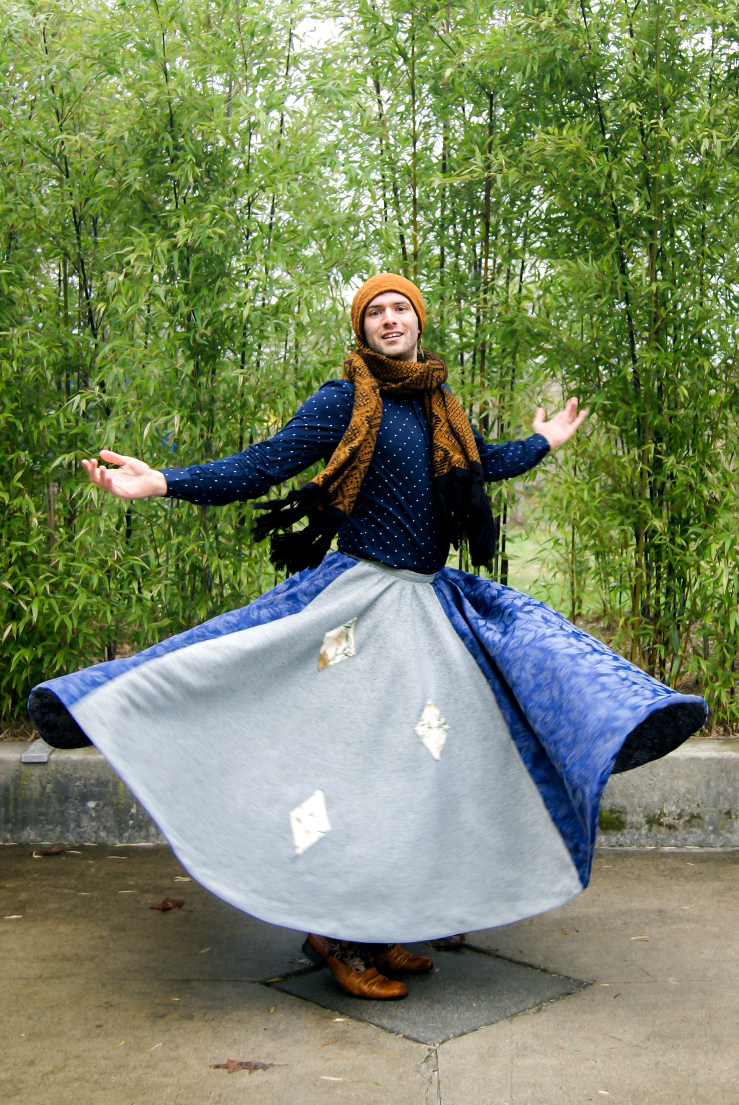 Image shows a tall, slightly bearded non binary person, wearing an orange-brown toque, a blue dress shirt, fringe earrings in blue, and a four paneled circle skirt made from repurposed curtain panels. They are spinning around and the skirt is in full movement, and they are facing the camera with their arms lifted to the sky, mid spin.