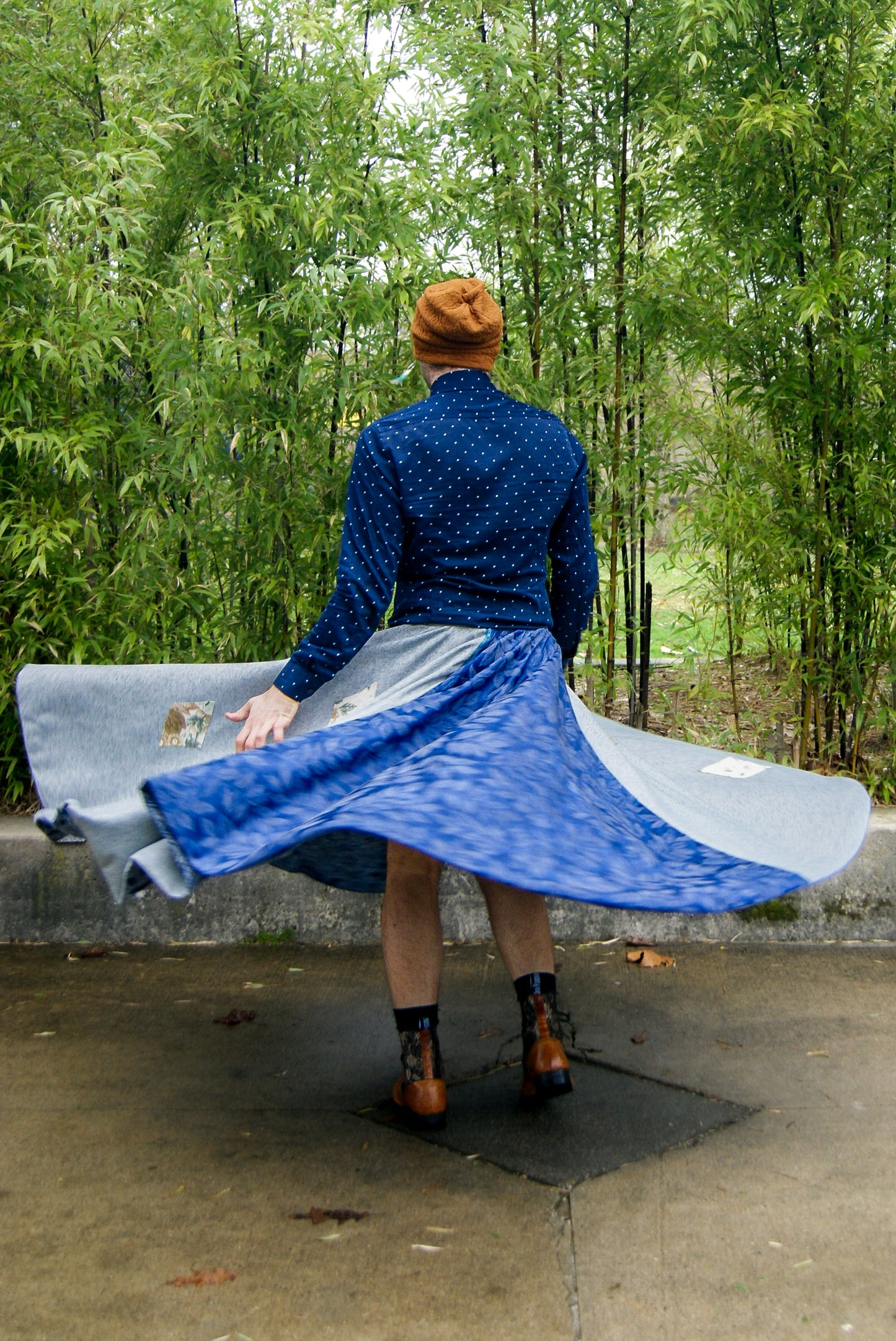 Image shows a tall, slightly bearded non binary person, wearing an orange-brown toque, a blue dress shirt, fringe earrings in blue, and a four paneled circle skirt made from repurposed curtain panels. They are spinning around and the skirt is in full movement, and their back is turned. 