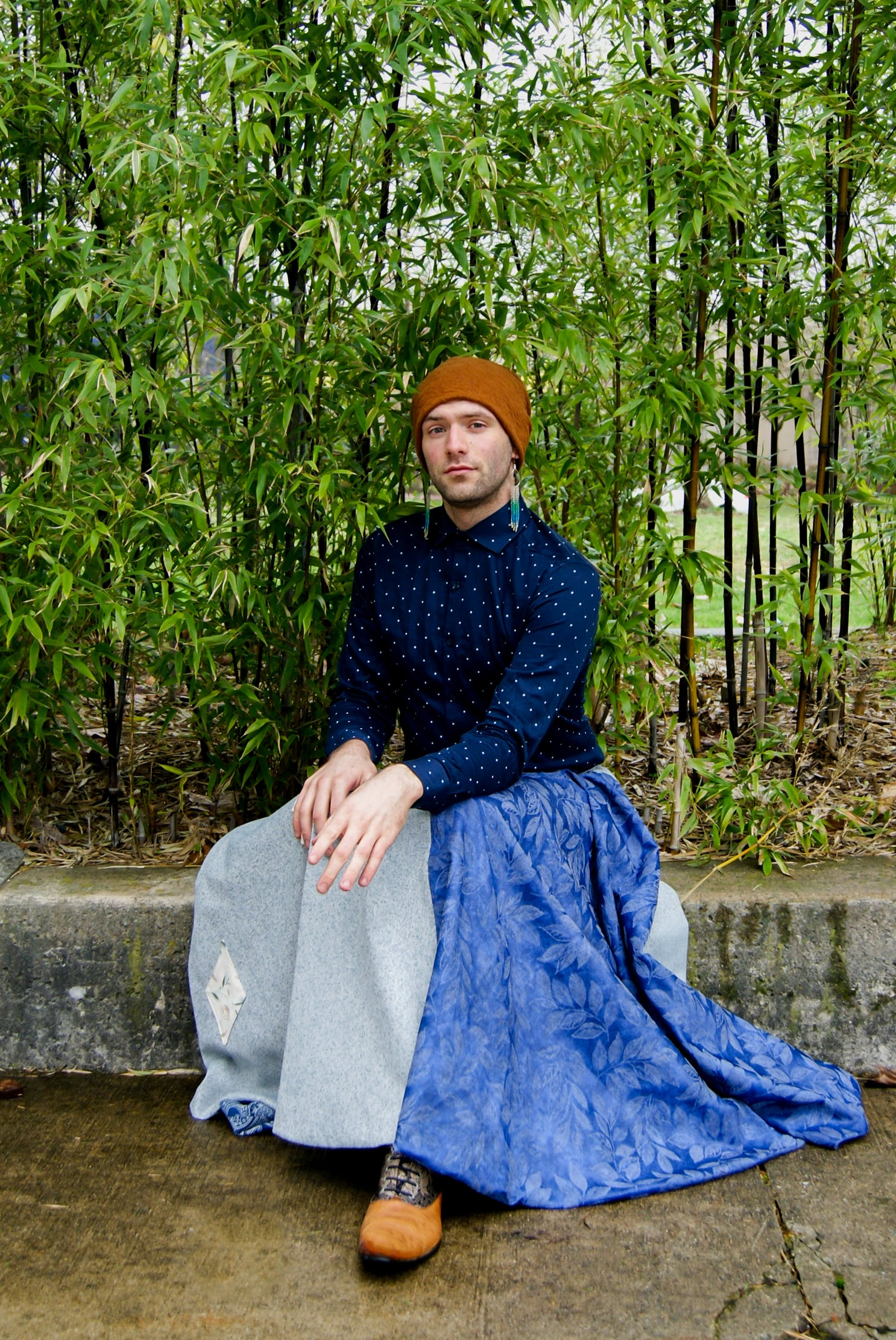 Image shows a tall, slightly bearded non binary person, wearing an orange-brown toque, a blue dress shirt, fringe earrings in blue, and a four paneled circle skirt made from repurposed curtain panels. They are seated elegantly on a short cement wall, in front of decorative bamboo, looking towards the camera, and their skirt falls around them on the cement