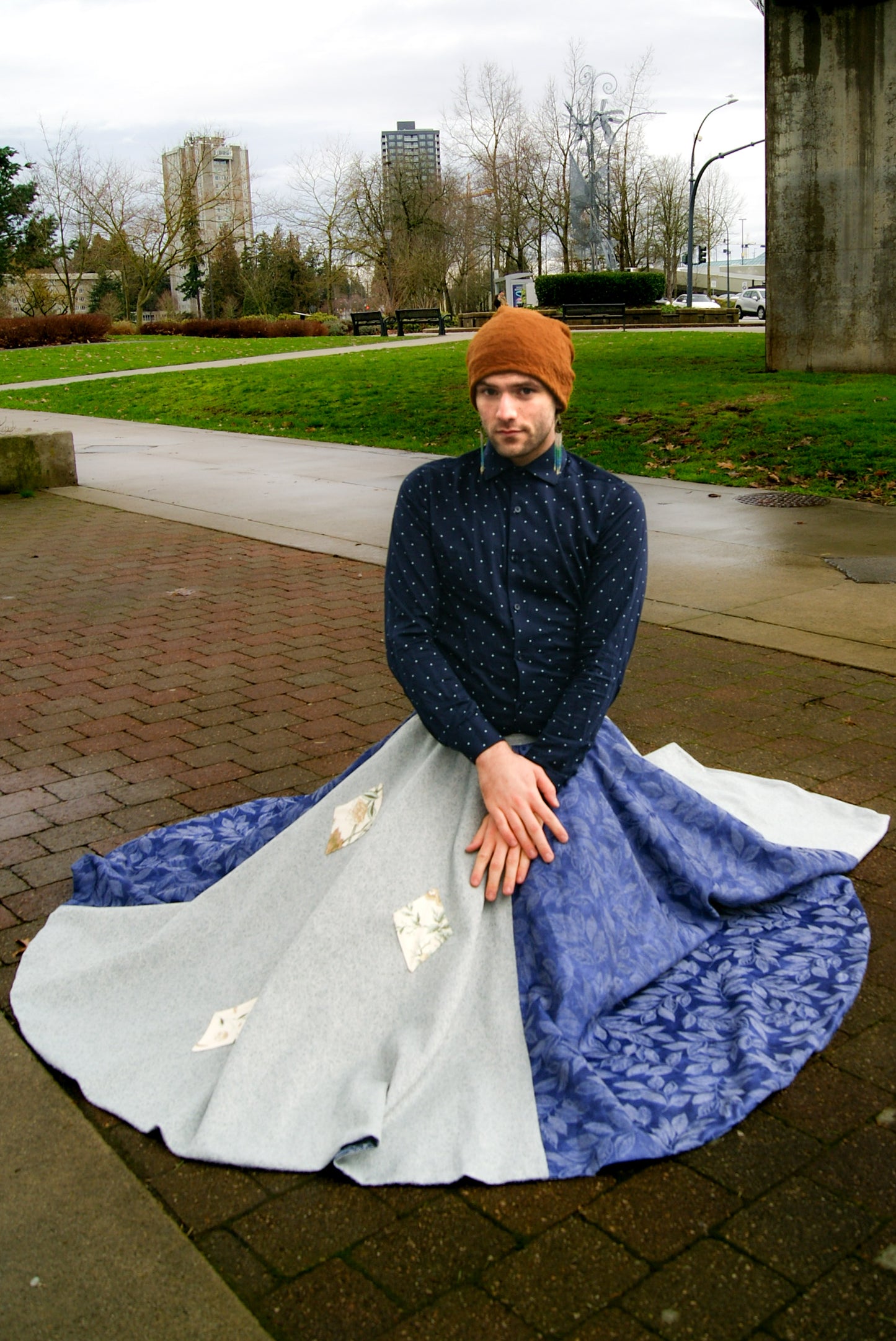 Image shows a tall, slightly bearded non binary person, wearing an orange-brown toque, a blue dress shirt, fringe earrings in blue, and a four panelled circle skirt made from repurposed curtain panels. They are perched on the ground, resting on their heels, and the skirt is laying around them on the brick, in a big circle