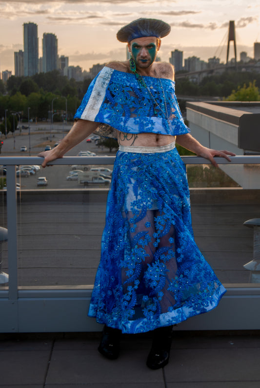 A person wearing a blue and white floral lace skirt with a flowing hem, paired with a blue top and black shoes, standing on a balcony with a cityscape in the background.