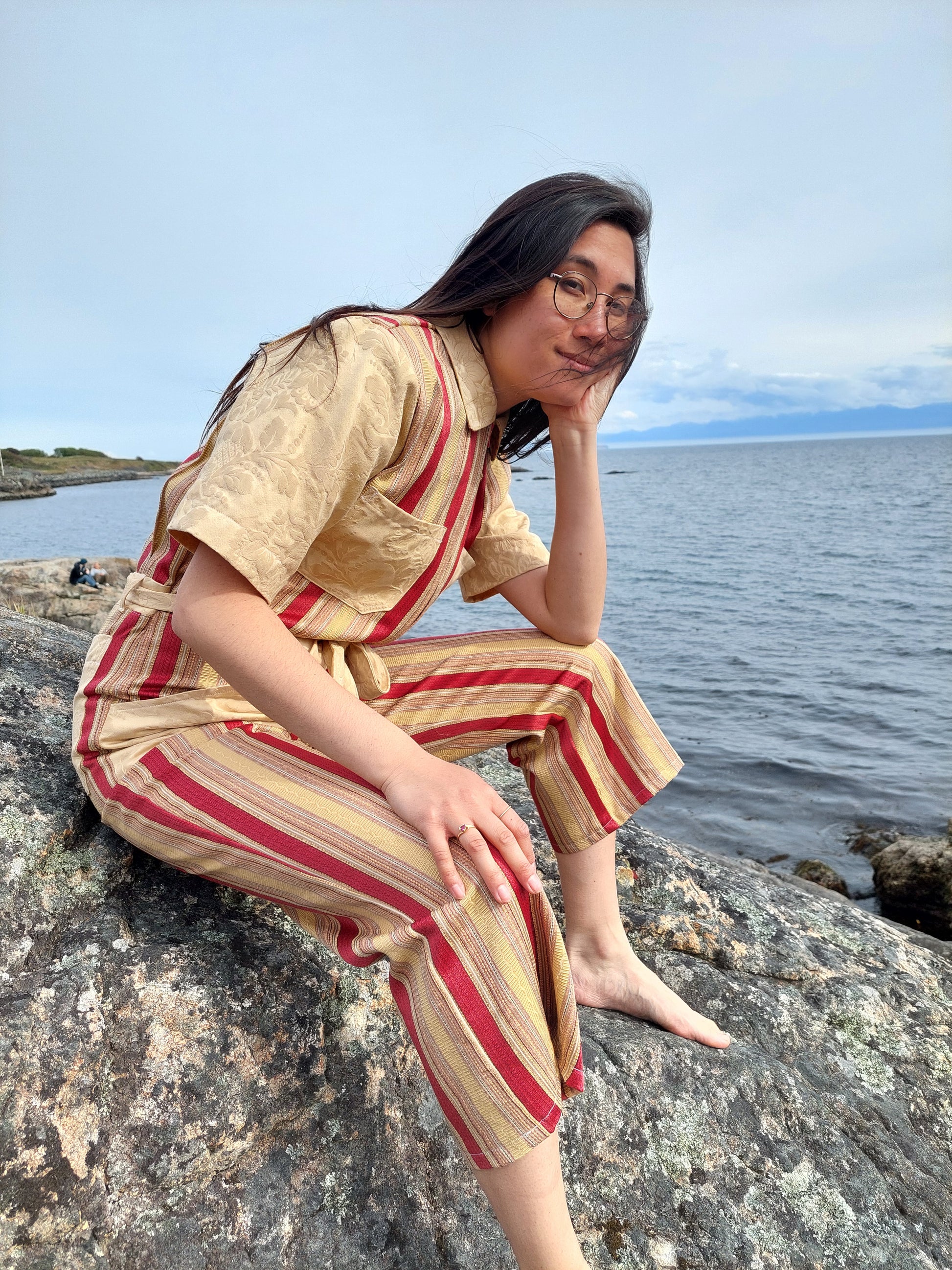 Model sits on the rocks at Saxe point, in Victoria BC. She looks towards the camera with a hand on her face, elbow planted on her knee. She wears a striped gold and red handmade jumpsuit she wears. She is smiling and  looks peaceful