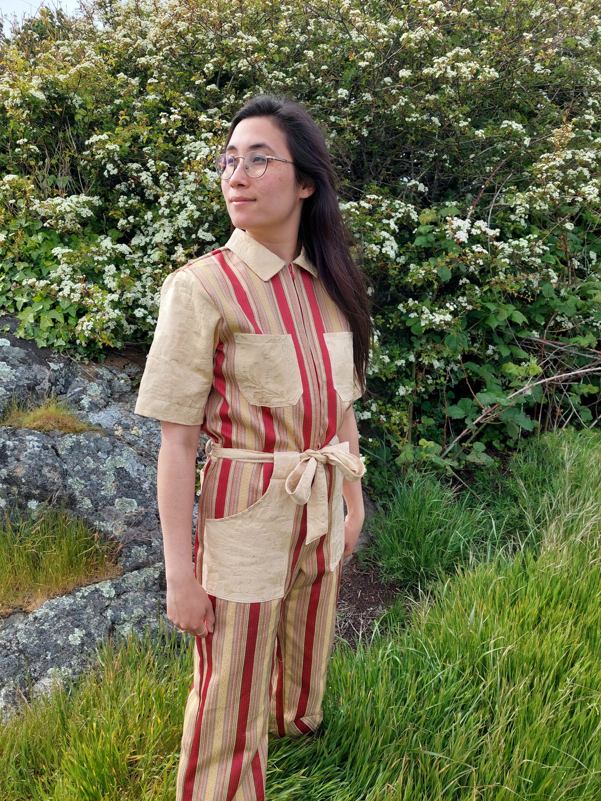 Model stands on in tall grass, in front of flowering bushes at Saxe point, in Victoria BC. She looks towards the ocean,  and her hair falls over the striped gold and red handmade jumpsuit she wears. She is smiling and  looks peaceful