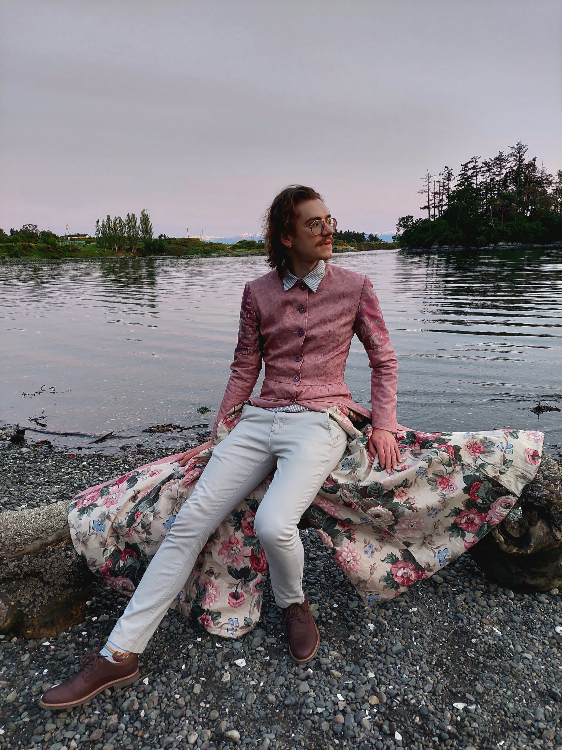 Model is seated on driftwood at a beach in Portage Park, in Victoria BC. The long coat they are wearing is draped on the driftwood done up in the front. The lining is a muted pink and green floral, and the coat is pastel pink, with lilac buttons on the front, and the cap of the sleeve is a lilac fabric as well. The model looks to the right, and the water behind them is tranquil.