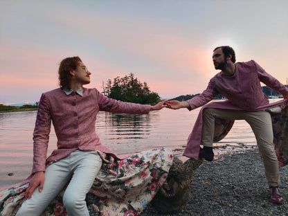 Two Models pose at a beach in Portage Park, in Victoria BC. The long coats they are wearing are a pastel pink, with lilac buttons and lilac sleeve accents. The lining is a muted pink and green floral, which shows at the bottom of the coat. One model is standing to the right, the person on the left is seated on a piece of driftwood. They look into each others eyes, reaching to touch hands, and the water behind them is tranquil.