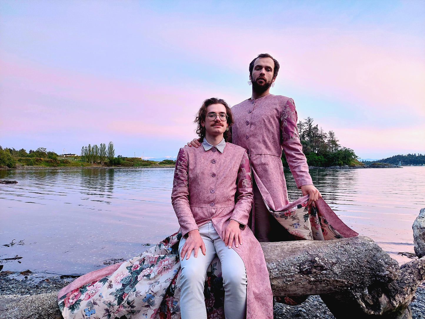 Two Models pose at a beach in Portage Park, in Victoria BC. The long coats they are wearing are a pastel pink, with lilac buttons and lilac sleeve accents.  The model on the left is seated on a piece of driftwood with the left side of their coat draped  beside them. The model on the right has their hand on the shoulder of the other model, and is standing behind the driftwood.