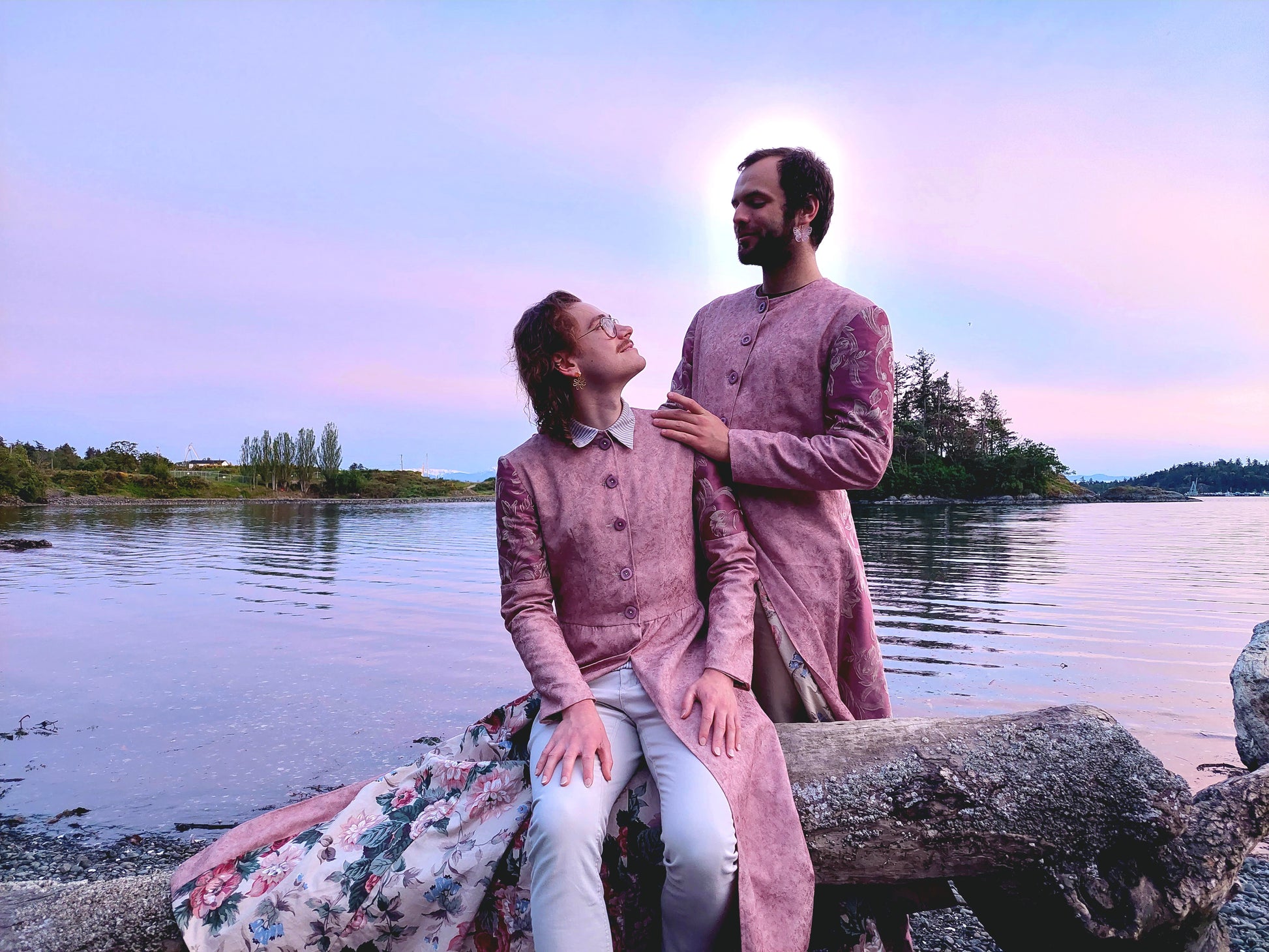 Two Models pose at a beach in Portage Park, in Victoria BC. The long coats they are wearing are a pastel pink, with lilac buttons and lilac sleeve accents.  The model on the right is standing and the model on the left is seated on a piece of driftwood. They are looking at each other with smiles