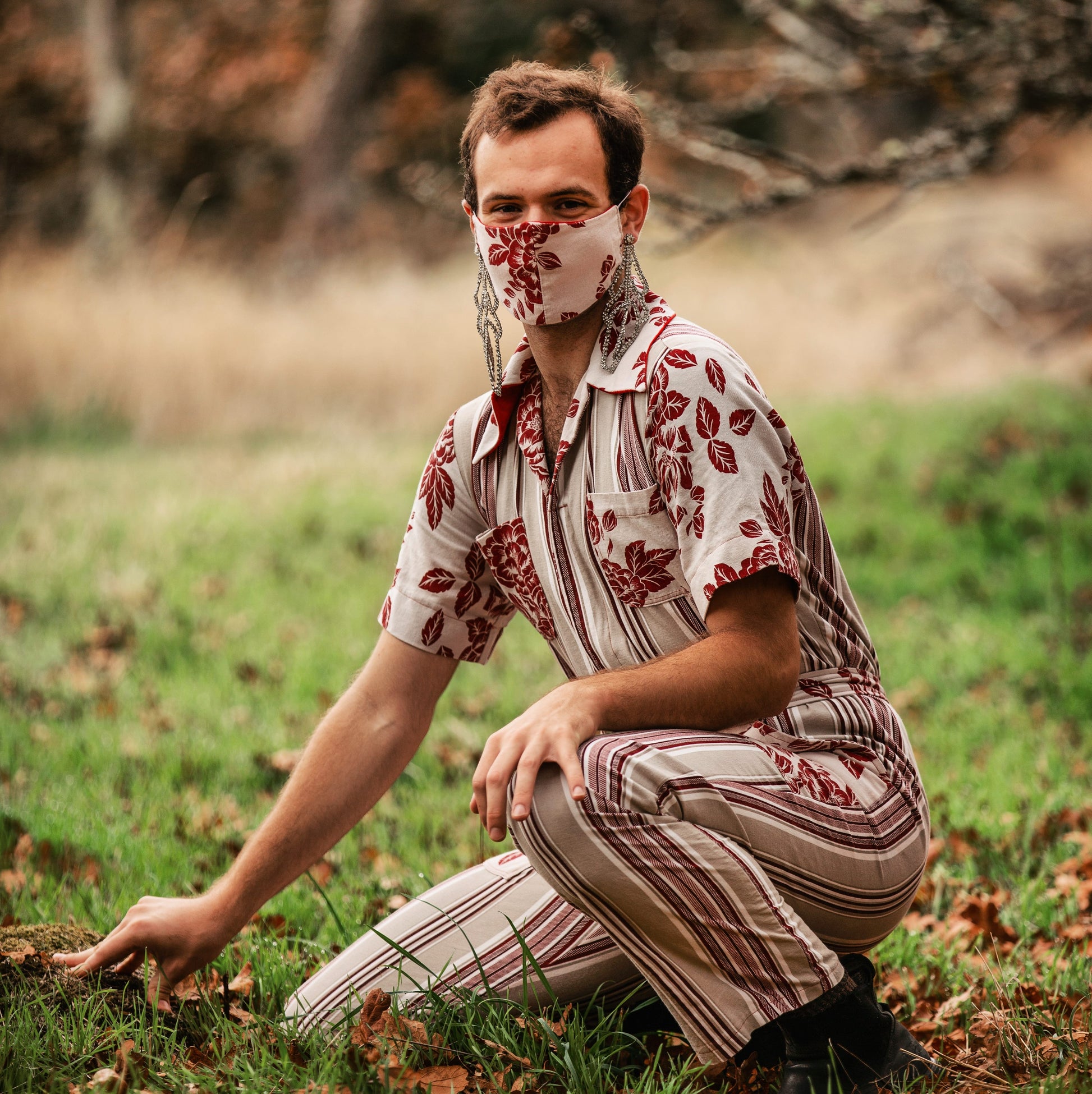 Model is wearing a red and beige vertically striped fitted jumpsuit and are crouching in a field of green grass. Behind them, oak branches cross the frame. They are wearing large dangling earrings and have a facemask which matches the accents on the jumpsuit, made from a fabric with red roses on it.