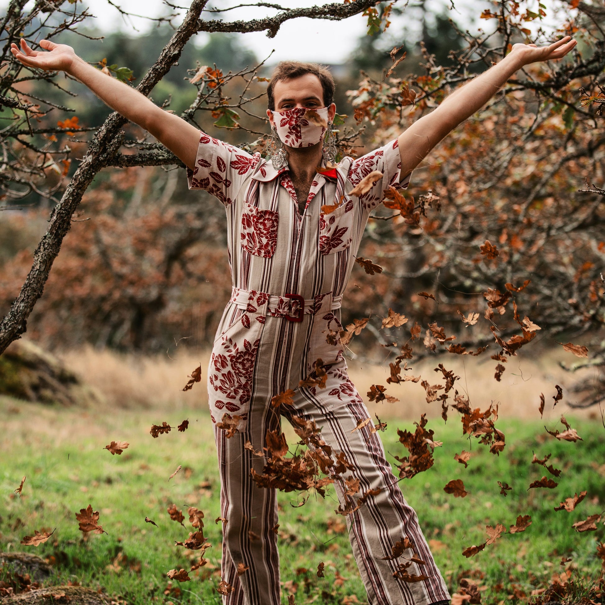 Model is wearing a red and beige vertically striped fitted jumpsuit and are standing in a field of green grass. Behind them, oak branches reach towards them and the model has their hands in the air as they have just tossed leaves into the air. They are wearing large dangling earrings and have a facemask which matches the accents on the jumpsuit, made from a fabric with red roses on it.